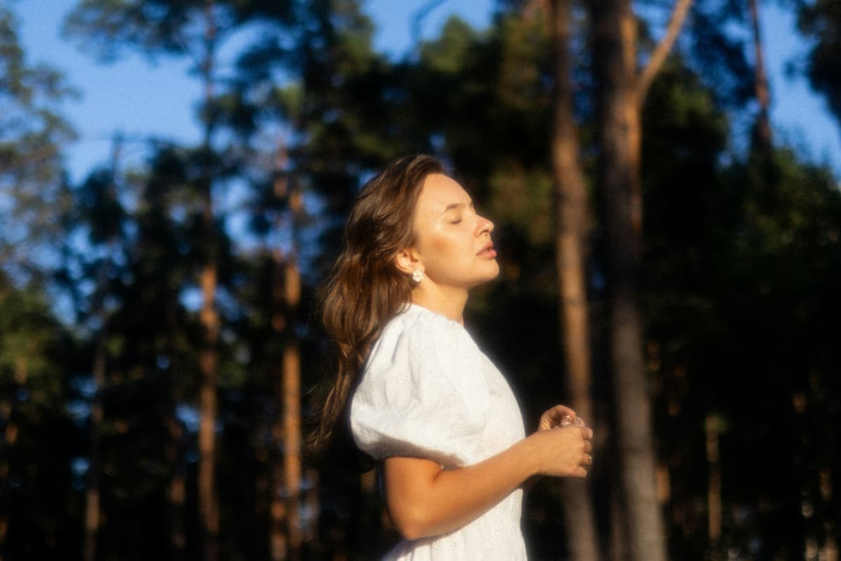 Fotografía de una mujer con los ojos cerrados y expresión de calma, vistiendo una camisa blanca en medio de un bosque rodeada de árboles. Representa el concepto de higiene del sistema nervioso, regulación del estrés y resiliencia frente al burnout biológico por SOMP.